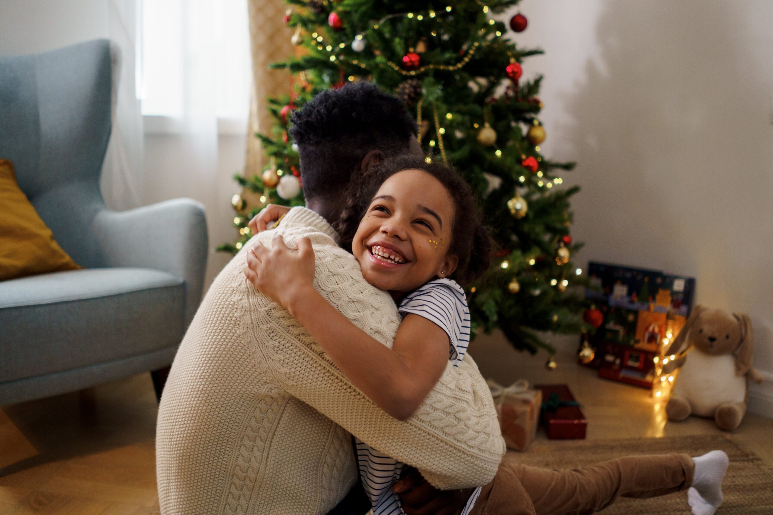 A kid hugging her dad in front of a Christmas tree.