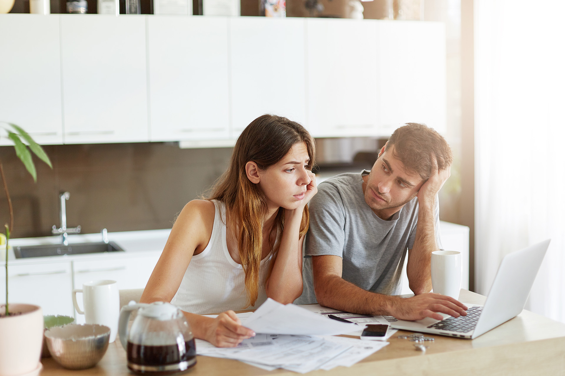 A worried young couple is reviewing their finances in the kitchen. They are surrounded by papers and a laptop, with the woman looking at documents and the man leaning his head on his hand, gazing at the laptop screen. A coffee pot, cup, and houseplant are also on the table, suggesting a domestic setting.