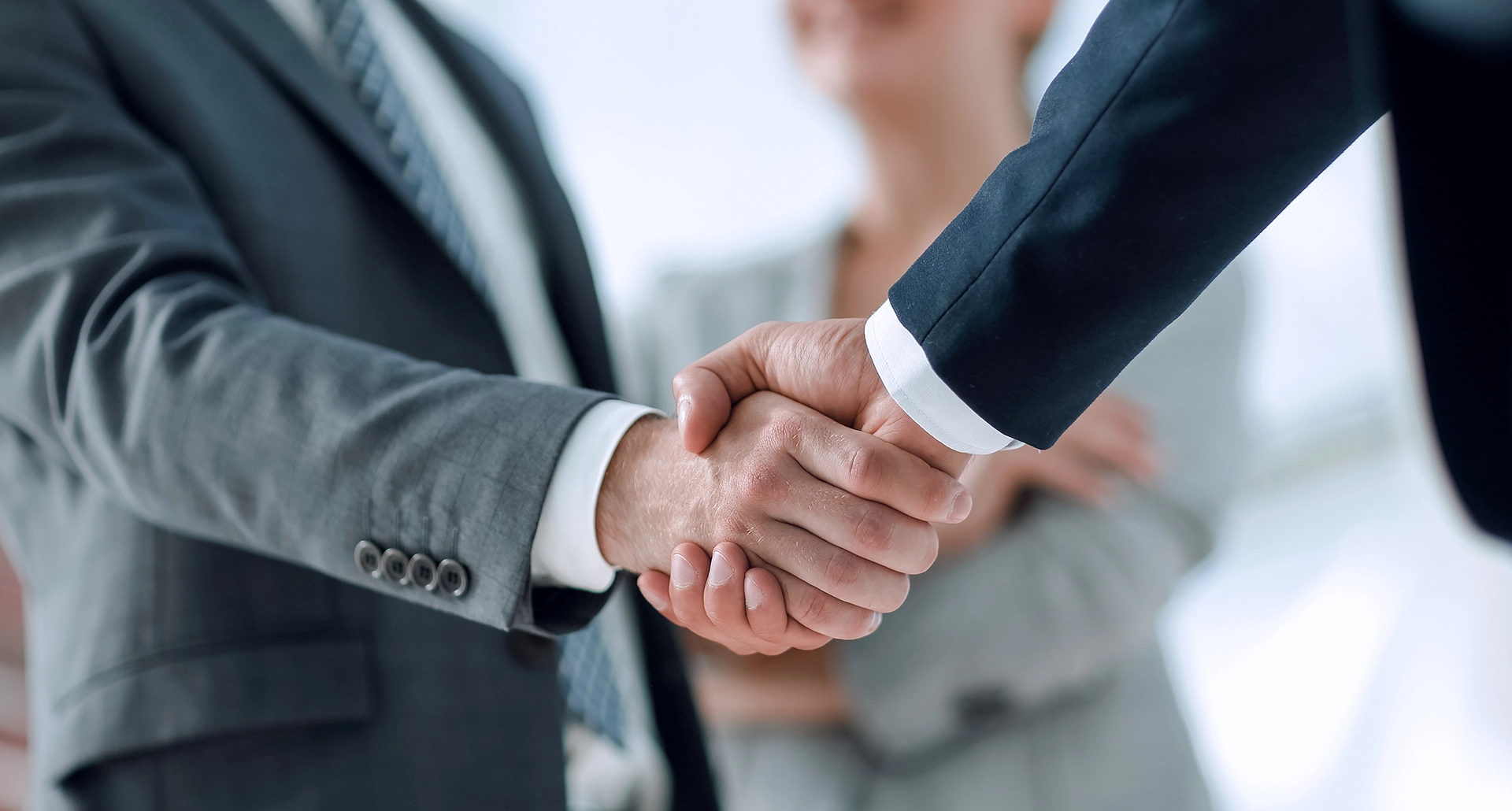 A close-up of two business professionals in suits shaking hands, with a smiling woman slightly blurred in the background.