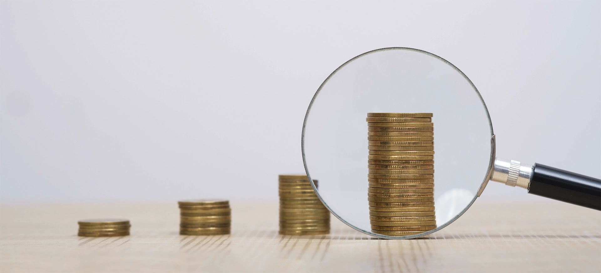 A magnifying glass focusing on a stack of gold coins, making them appear larger, while smaller stacks of coins in the background gradually increase in height